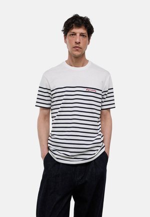 Homme aux cheveux bouclés foncés portant un t-shirt à manches courtes rayé blanc et noir avec un texte rouge "Legendaddy" et un pantalon foncé, mains dans les poches.