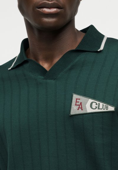 Ciemnozielona prążkowana koszulka polo z kołnierzykiem wykończonym białym paskiem i trójkątną naszywką z napisem "EA CLUB" na lewej piersi.