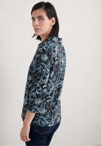 Blusa nera con un pattern floreale blu e accenti verdi, caratterizzata da maniche lunghe e colletto. Il materiale appare texturizzato e leggero.