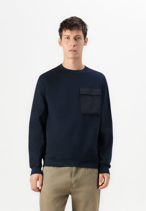 Marinefarbener Sweatshirt aus Baumwollmischung, mit Fronttasche und Klappe, gerippten Bündchen und Rundhalsausschnitt. Getragen mit beigen Hosen.