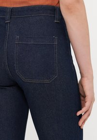 Detailaufnahme einer Person, die eine dunkelblaue Jeans trägt, mit sichtbarem Gesäßtasche und einer Hand, die entspannt an ihrer Seite liegt.