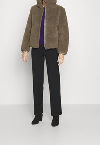 Hnědá fleecová bunda se zipem a kapucí, s elastickými manžetami, kombinovaná s černými kalhotami a kotníkovými botami. Látka působí měkce a strukturovaně.