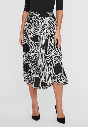 Kobieta w czarno-białej, abstrakcyjnej spódnicy midi w wzór zebry, czarne szpilki z zaostrzonym noskiem oraz czarna bluzka z rękawami do połowy ramienia.