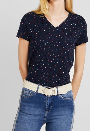 Marineblå V-hals kortærmet skjorte med flerfarvet småprikket mønster, båret sammen med blå jeans og hvidt bælte med rund spænde.