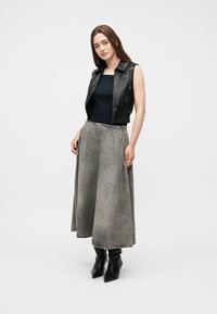 Γυναίκα με μαύρο αμάνικο δερμάτινο γιλέκο, μαύρο τοπ, μακριά γκρι υφασμάτινη φούστα και μαύρες μυτερές μπότες, που στέκεται μπροστά σε απλό φόντο.