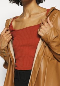 Béžový kabát z umělé kůže s béžovou podšívkou a knoflíkovými detaily, vrstvený přes žebrovaný červený top. Bez viditelných vzorů nebo grafik.