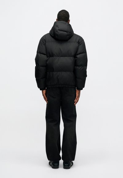 Melnā pūkainā jaka ar kapuci, ar kvadrātu dizainu un elastīgu apakšmalu, pārī ar tumšu denimu biksēm. Minimalistisks un vienkāršs izskats.