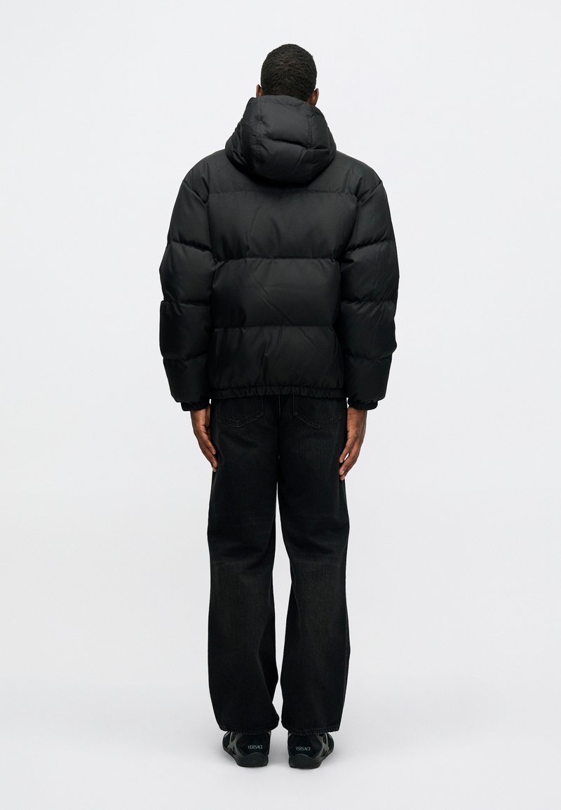 Sort pufferjakke med hætte, med et quiltet design og elastisk kant, parret med mørke denimbukser. Minimalistisk og strømlinet udseende.