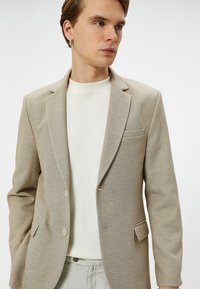Beige strukturerad blazer med två knappar, slagkrage och fickor med klaff, dragen över en ljusfärgad rundhalsad tröja.