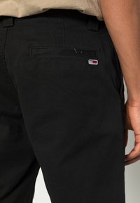 Černé bavlněné kalhoty s zadní kapsou a malým vyšitým logem. Materiál má hladkou texturu a klasický střih.