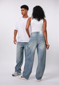 Lyseblå baggy denimjeans med en falmet tekstur og detaljert sidepute, kombinert med en hvit tanktop og en overdimensjonert hvit T-skjorte.