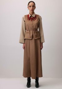 Barna velúr mellény, övvel, egy bézs blúzra rétegezve, egy hozzá illő barna maxi szoknyával és fekete, hegyes orrú csizmával párosítva.