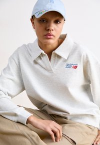 Svetlosivá polo mikina s veľkým golierom a vyšitým logom "New Balance" v modrej a červenej farbe. Nosí sa s béžovými nohavicami. Svetlomodrá čiapka.