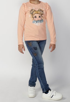 Niño con camisa de manga larga color durazno con estampado de niña caricatura, jeans azules con bordado de mariposas y zapatillas blancas con tiras de velcro.