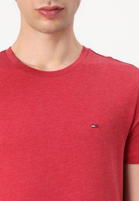 Rotes T-Shirt mit kurzen Ärmeln aus weichem, strukturiertem Stoff. Enthält ein kleines, gesticktes Logo in Blau, Weiß und Rot auf der linken Brustseite.