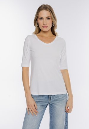 Blondynka w dużych kolczykach, białej koszuli z rękawami do łokcia i jasnoniebieskich dżinsach, stojąca na jednolitym białym tle.