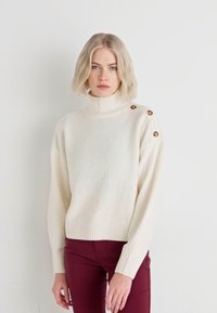 Suéter de cuello alto crema con puños acanalados, botones decorativos en el hombro y un ajuste relajado. Lleva pantalones burdeos.