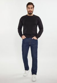 Czarny prążkowany sweter, niebieskie dżinsy i białe trampki. Model stoi z rękami w kieszeniach na jasnym tle. Prosta i casualowa stylizacja.