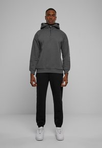 Mørkegrå hættetrøje med lynlås ved halsen, parret med sorte sweatpants og hvide sneakers. Hættetrøjen har en stor hætte og ribbede manchetter.