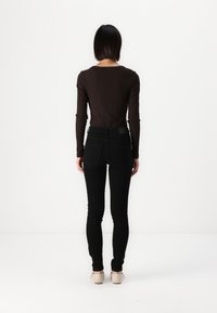 Καφέ ριγωτό τοπ με μακριά μανίκια συνδυασμένο με μαύρα στενά τζιν με πίσω τσέπες. Το μοντέλο φοράει ανοιχτόχρωμα παπούτσια. Χαλαρό φόντο.