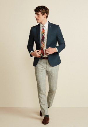 Marineblazer over een wit shirt, gecombineerd met een grijs geruit broek en een multicolore stropdas. Bruine loafers maken de outfit compleet.