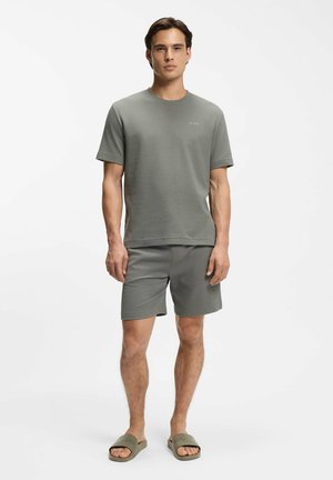 Uomo che indossa una maglietta a maniche corte e pantaloncini grigi con trama coordinata e sandali verdi, in piedi su uno sfondo bianco uniforme.