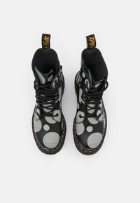 Botas negras con una superficie brillante, grandes lunares blancos, rayas horizontales, cordones negros y costuras amarillas a lo largo de la suela.