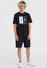 Schwarzes Baumwoll-T-Shirt mit grafischem Druck, kombiniert mit passenden schwarzen Shorts. Weiße Sneakers mit schlichtem Design und Schnürsenkeln.