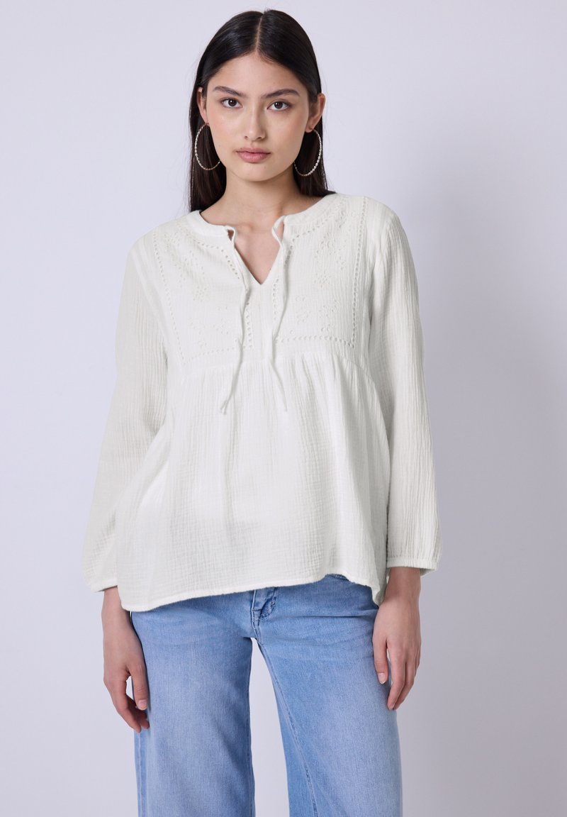 Hvid struktureret bluse med lange ærmer, V-hals og dekorativ syning. Kombineret med lyseblå jeans.