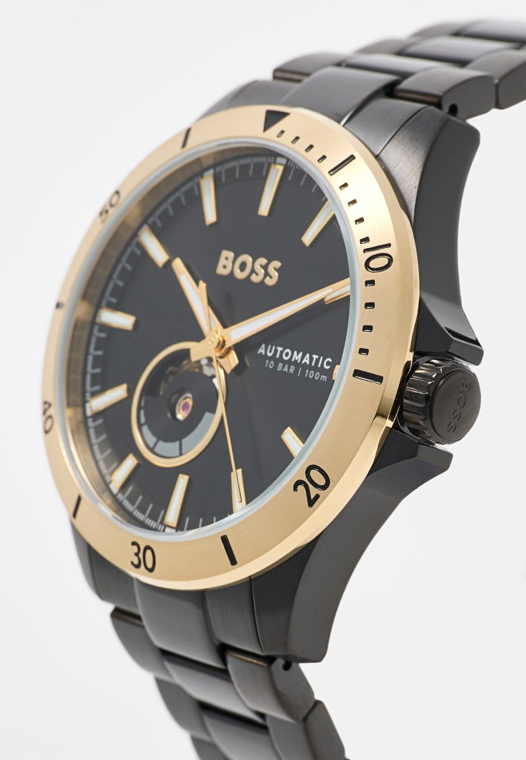 Montre automatique avec un cadran noir, des accents dorés et un bracelet en métal noir. Elle présente un mouvement visible et une étanchéité jusqu'à 100 m.