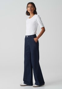 Witte short-sleeve top gecombineerd met hoge taille, wijde broekspijpen van donkerdenim. Zakdetails en klassieke knoopsluiting. Casual schoenen maken de look compleet.