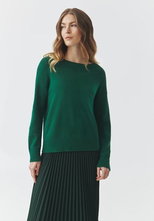 Green knit sweater with a rounded neckline and ribbed hem, paired with a pleated dark green skirt. Soft texture and relaxed fit.