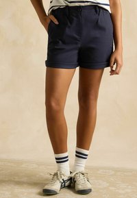 Marineblaue Baumwollshorts mit umgeschlagenem Saum, ausgestattet mit einem Knopfverschluss und Seitentaschen, getragen mit weißen Socken und pastellfarbenen Sportschuhen.