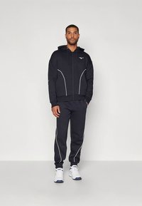 Treino desportivo azul escuro com capuz com fecho e detalhes brancos, combinado com calças a condizer. Tecido texturizado; combinado com ténis brancos.