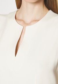 Blouse couleur crème avec un décolleté en V, en tissu lisse et avec des détails de couture subtils. Design simple avec une coupe décontractée.