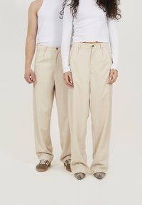 Pantalons beiges taille haute avec un design plissé, devant plat et passants de ceinture. Portés avec des tops blancs et des chaussures variées. Texture lisse.