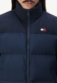 Mornarska pufasta jakna z ribastim visokim ovratnikom, zadrgo in oblazinjenim videzom. Ima majhen logo v rdeči, beli in modri barvi na prsih.