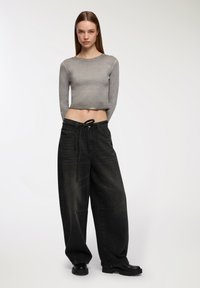 Młoda kobieta z długimi prostymi włosami, ubrana w szary krótki rękaw, luźne czarne dżinsy i czarne botki, stojąca na białym tle.