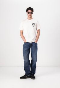 White short-sleeve t-shirt with "NYC" logo, blue denim jeans, and black sneakers. The fabric appears smooth; jeans show slight distressing.