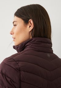 Giacca puffer bordeaux con colletto alto, caratterizzata da un design a rombi trapuntati e un logo discreto vicino al collo. Texture liscia e lucida.