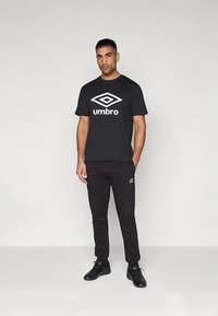 Melnā īsroku krekls ar baltu Umbro logo, kombinēts ar melnām sporta biksēm ar mazu logotipu uz augšstilba, un melnām sporta apaviem.