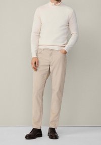 Homme portant un pantalon chino beige, un pull en maille crème par-dessus une chemise à col, et des chaussures habillées en cuir marron foncé, debout contre un mur clair uni.