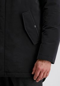 Zwarte pufferjack met een gladde afwerking, voorzien van een zijzak en een manchet mouw. Inclusief een subtiele gestreepte accent op de zak.