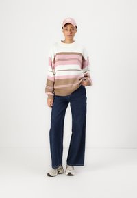 Striped knit sweater in cream, pink, and brown. Paired with wide-leg dark denim jeans and beige sneakers. Pink baseball cap accessory.