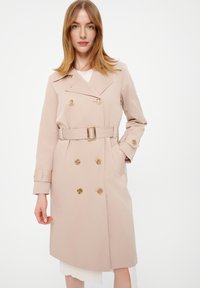 Beżowy płaszcz trench z podwójnym zapięciem, złotymi guzikami, paskiem w talii oraz długimi rękawami z zapięciem na guziki.