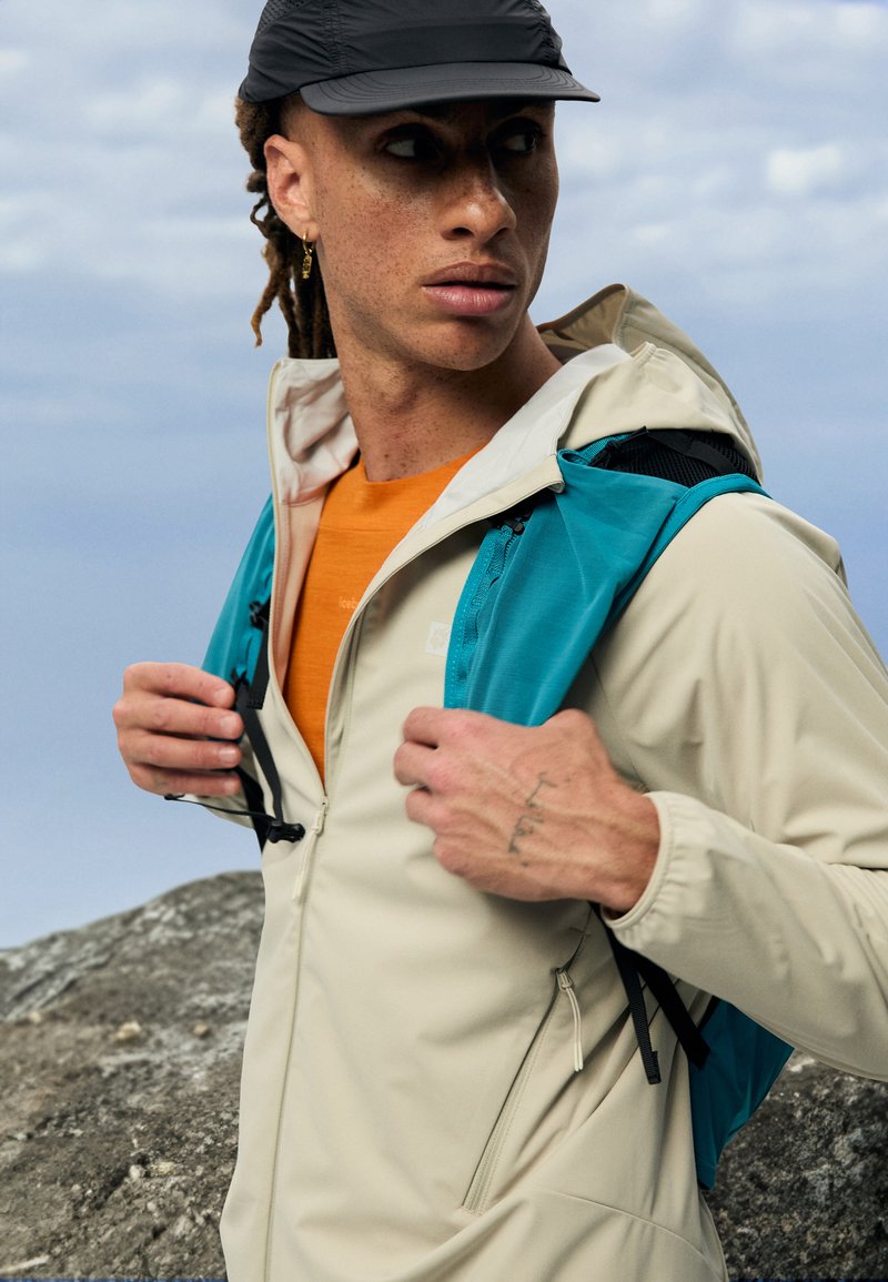 Veste imperméable beige clair avec capuche et poches zippées, superposée sur une chemise orange, accompagnée d'un sac à dos teal sur l'épaule.