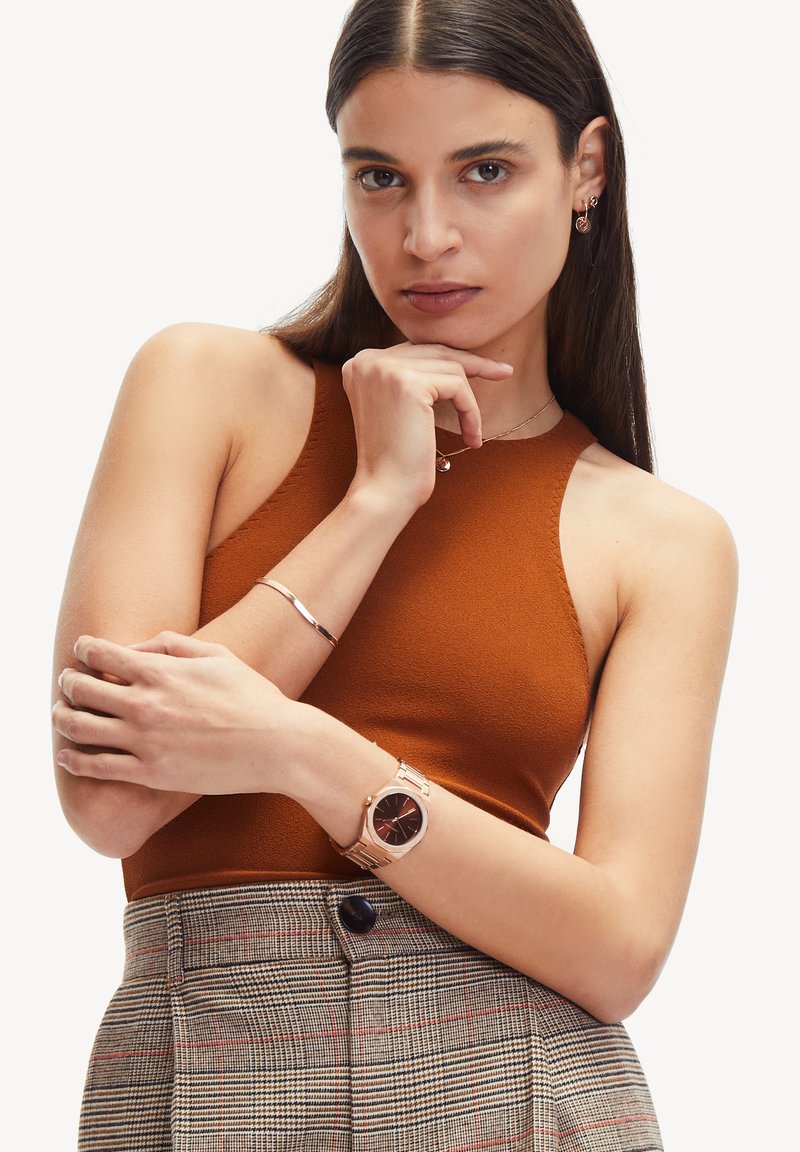 Bronzen mouwloos topje gecombineerd met een geruite hoge taille rok. Roze gouden horloge met een donkere wijzerplaat en een eenvoudige band. Zilveren armband.