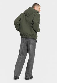 Oliwkowa, kapturowa kurtka z elastycznym w pasie, zestawiona z szarymi dżinsami denimi z tylnymi kieszeniami i delikatnym wyblaknięciem. Całość dopełniają czarne sneakersy.