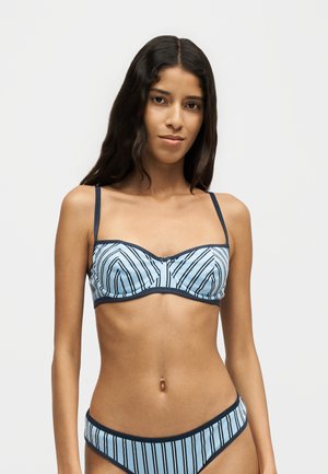 Donna che indossa un bikini a righe azzurro chiaro e blu navy con spalline sottili, in piedi davanti a uno sfondo grigio chiaro uniforme.
