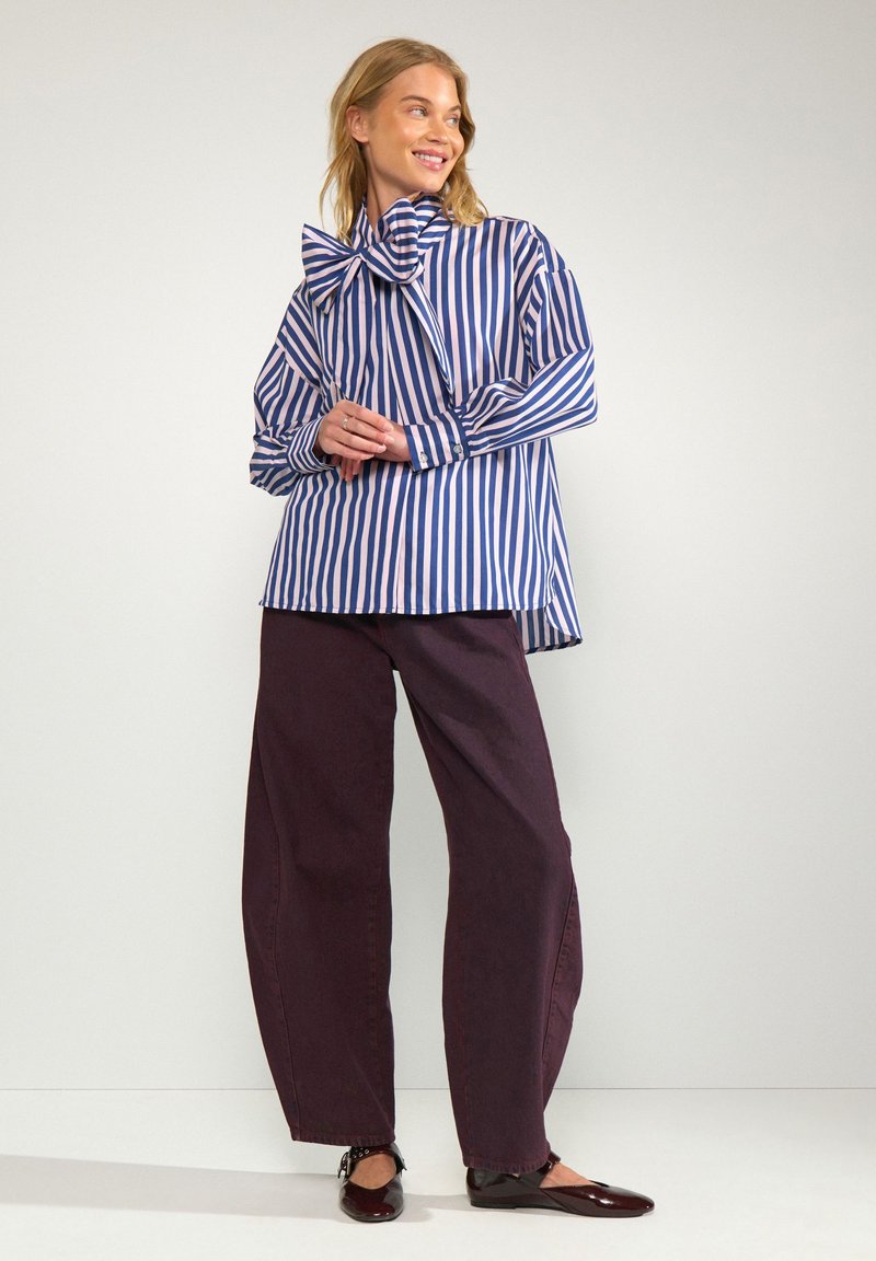 Femme souriante debout portant une chemise à rayures bleu et blanc avec un grand nœud et un pantalon large foncé avec des chaussures plates.